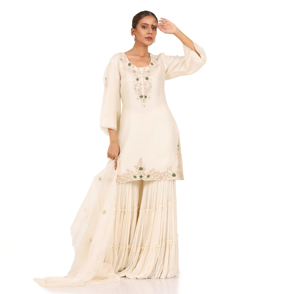 A woman standing and wearing Off White Kurta Sharara Set.