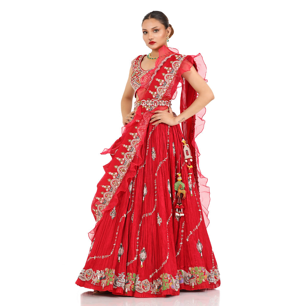 A woman standing and wearing Coral Red Lehenga Set with a waist belt attaining a bridal attire.