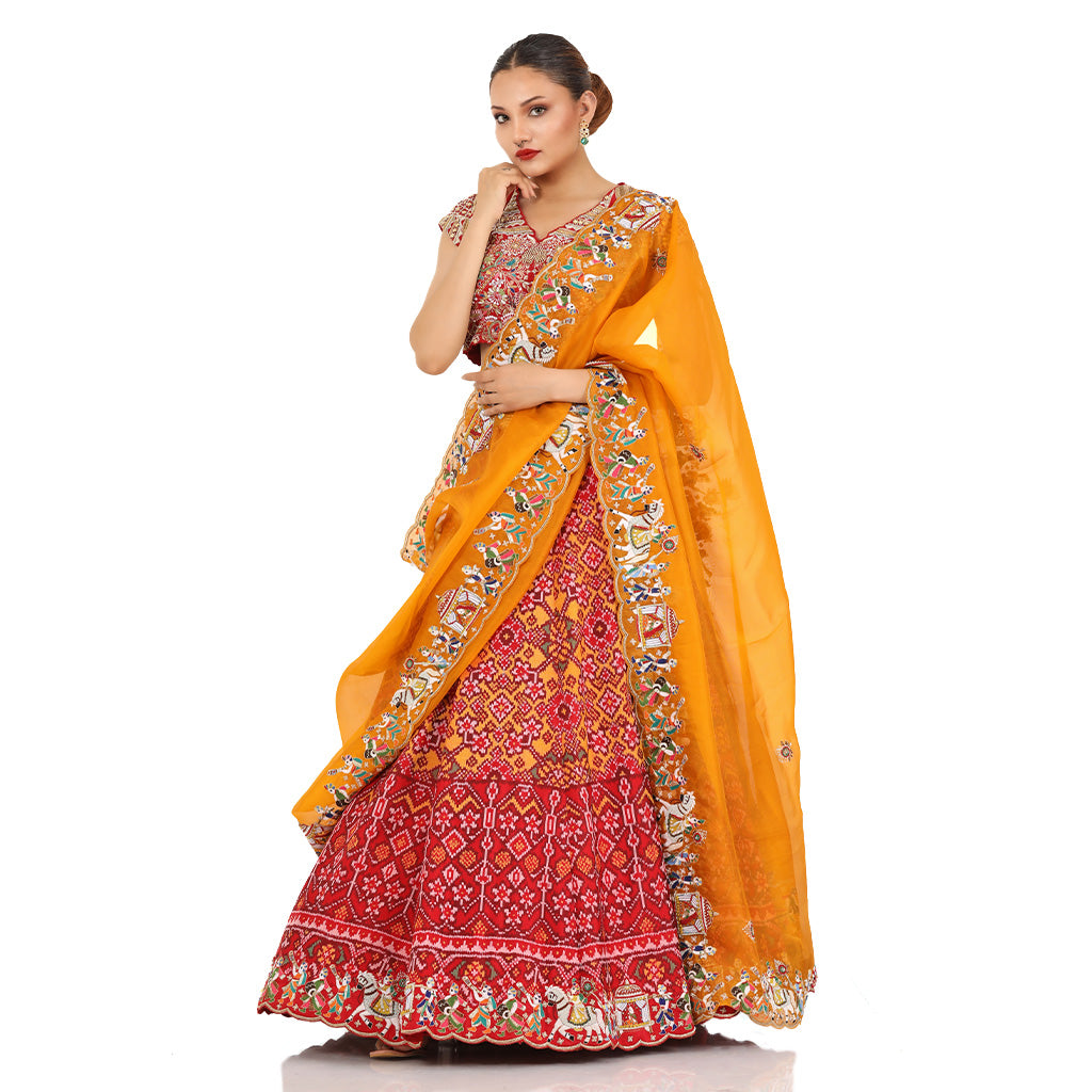 A woman standing and wearing Red orange Patola lehenga attaining a bridal attire.