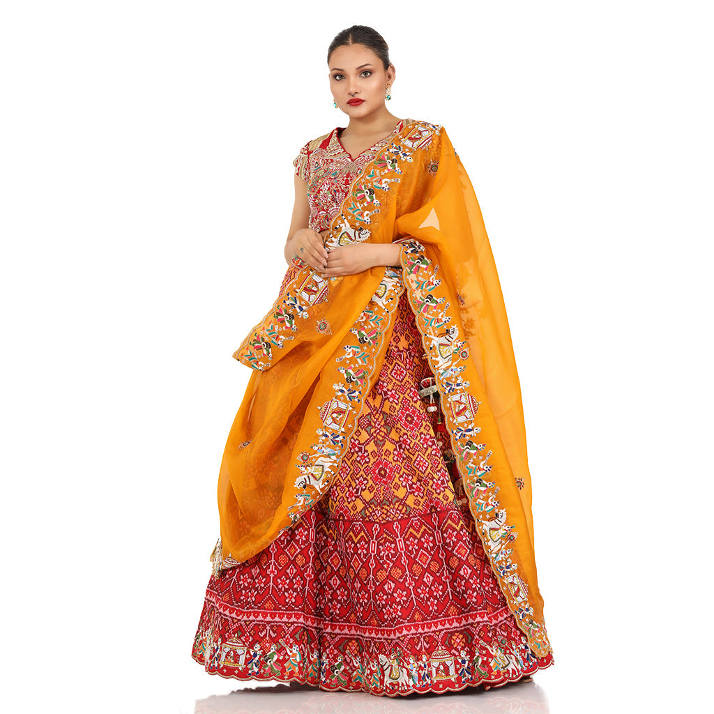 A woman standing and wearing Red orange Patola lehenga attaining a bridal attire.