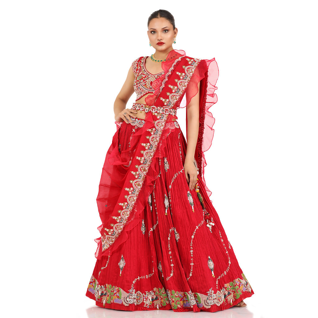 A woman standing and wearing Coral Red Lehenga Set with a waist belt attaining a bridal attire.