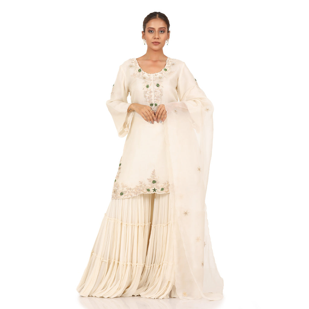 A woman standing and wearing Off White Kurta Sharara Set.