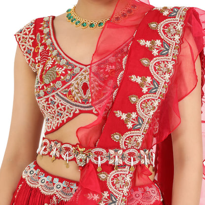 A woman standing and wearing Coral Red Lehenga Set with a waist belt attaining a bridal attire.