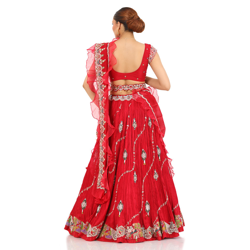 A woman standing and wearing Coral Red Lehenga Set with a waist belt attaining a bridal attire.