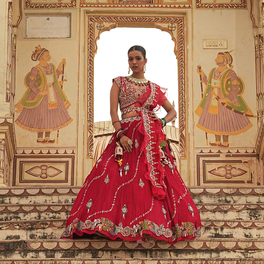 A woman standing and wearing Coral Red Lehenga Set with a waist belt attaining a bridal attire.