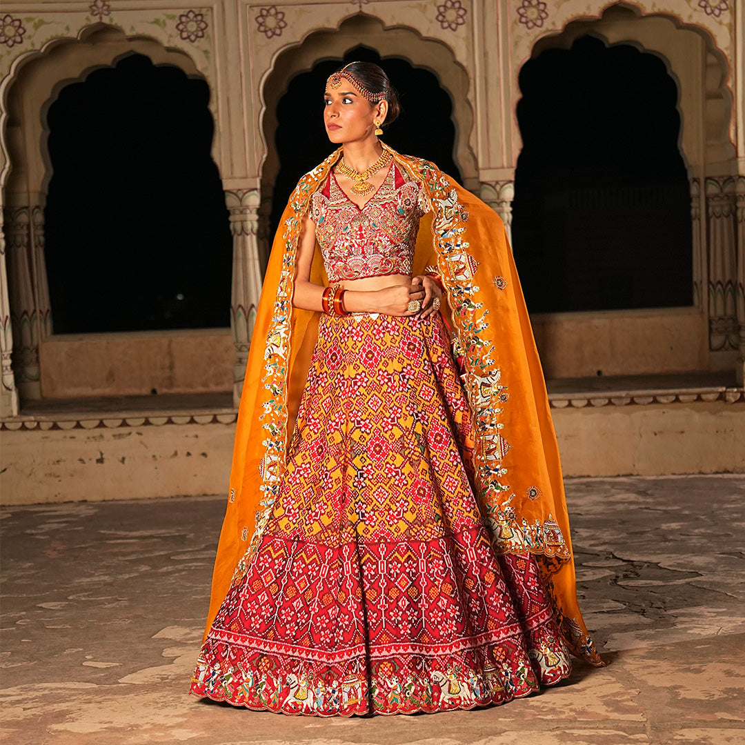 A woman standing and wearing Red orange Patola lehenga attaining a bridal attire.