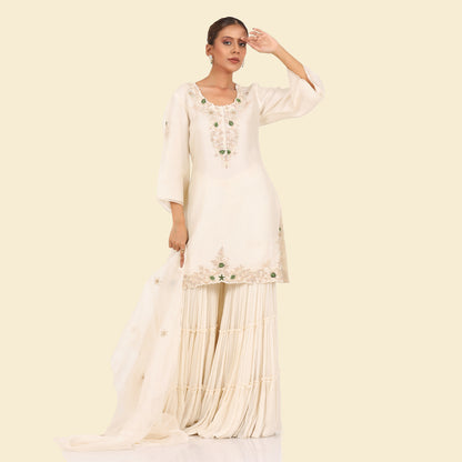 A woman standing and wearing Off White Kurta Sharara Set.